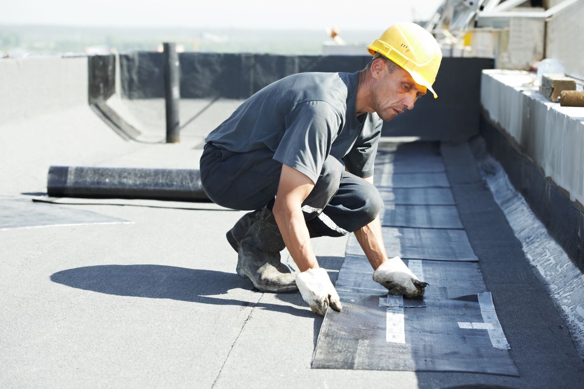 A construction worker in a yellow hard hat lays black roofing material on a flat rooftop under clear skies.