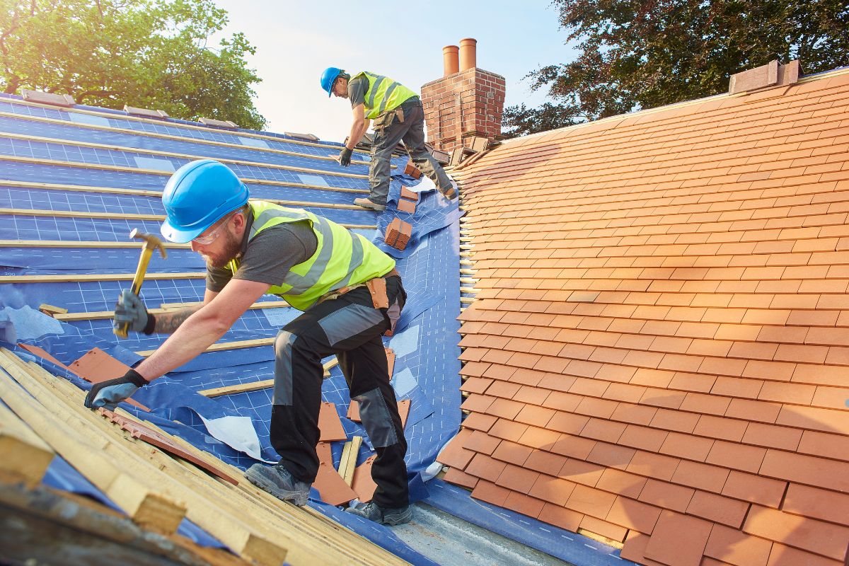 Two workers in helmets and safety vests are installing roof tiles on a sloped roof under clear blue skies.