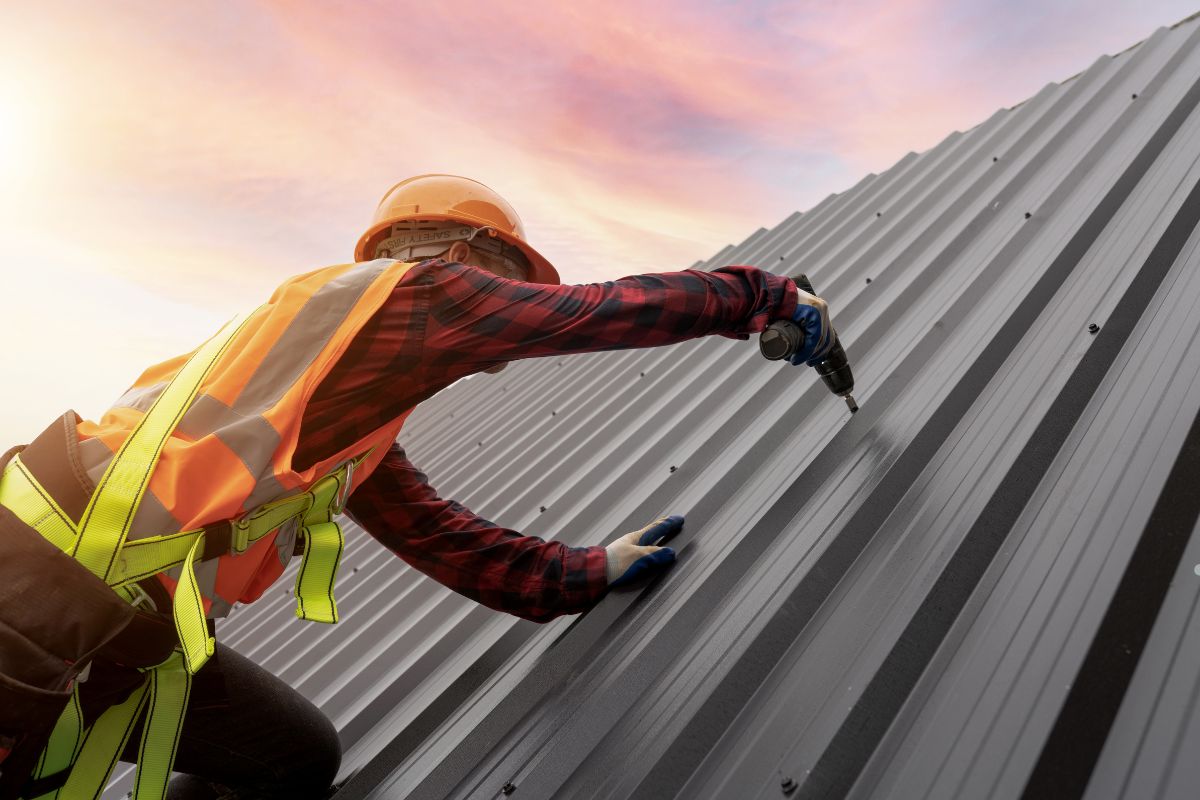A construction worker in a safety vest and helmet drills into a metal roof at sunset, demonstrating roofing expertise.